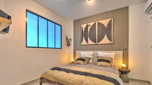 A modern bedroom with a large bed, a window, a wall-mounted artwork, and a bedside table with a lamp. - Apartment TalyJay - 2 bedrooms with swimming pools (Nettle Bay)