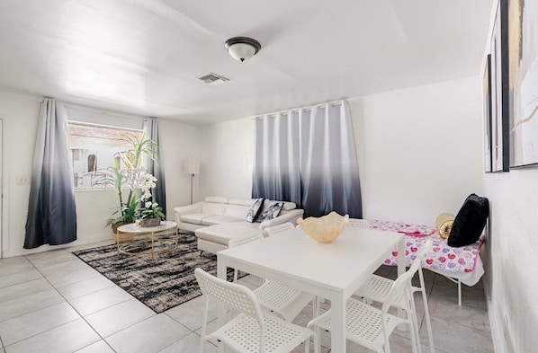 A modern living room with a white sofa, a dining table with chairs, a small coffee table, and a plant.