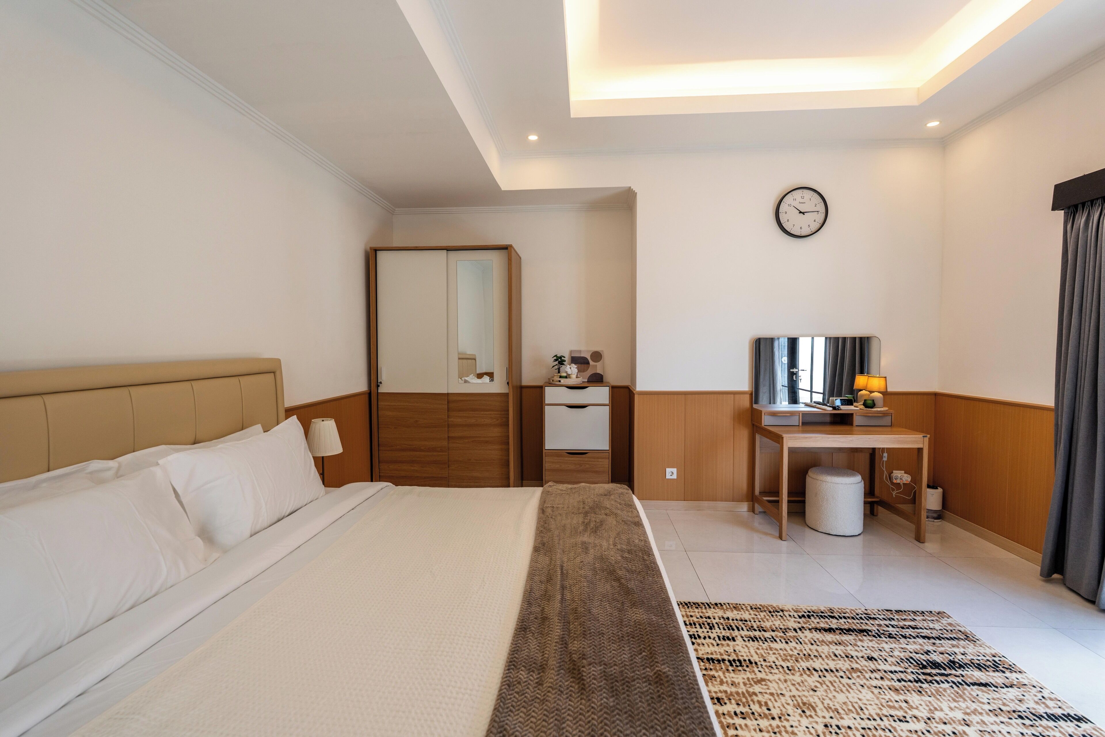 A hotel room with a bed, a desk, a mirror, a clock, and a window with curtains.