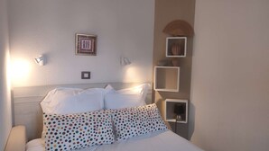 A neatly made bed with white bedding and colorful polka dot pillows, a framed picture on the wall, and a wall-mounted fan. - Charmant T2 vue mer pour deux avec piscine (ARZON)