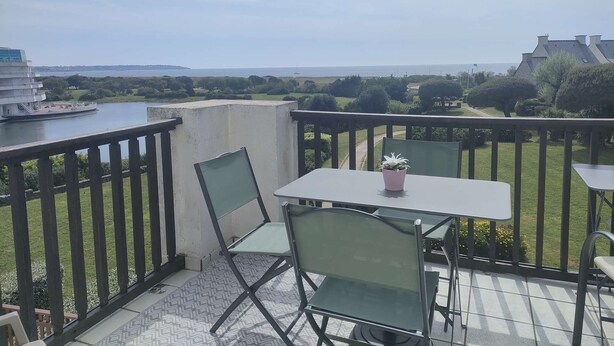 A balcony with a table and chairs, overlooking a body of water and greenery. - Charmant T2 vue mer pour deux avec piscine (ARZON)
