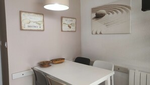 A dining area with a white table, two chairs, a hanging light fixture, and wall art depicting a beach scene. - Charmant T2 vue mer pour deux avec piscine (ARZON)