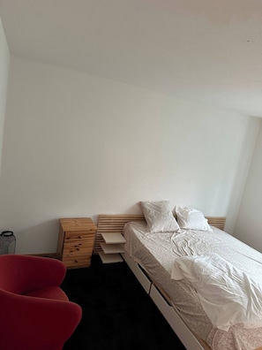 A bedroom with a bed, a red chair, and a wooden nightstand with drawers. - Room-a Furnished Room in our Cozy Apart (Baden)