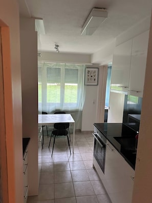 A compact kitchen with a dining table, chairs, a black stove, and a window with blinds. - Room-a Furnished Room in our Cozy Apart (Baden)