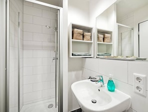 A bathroom with white tiled walls, a glass shower enclosure, a white sink with a faucet, and shelves with baskets. - Room-a Furnished Room in our Cozy Apart (Baden)