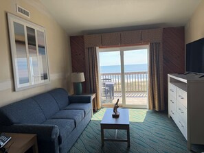 Une chambre d’hôtel avec un canapé, une petite table avec une lampe, une télévision, un balcon donnant sur la plage et une moquette à motifs.