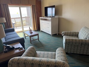 Quarto de hotel com sofá, poltrona, mesa pequena com objeto decorativo, televisão em móvel e porta de vidro deslizante para uma varanda.