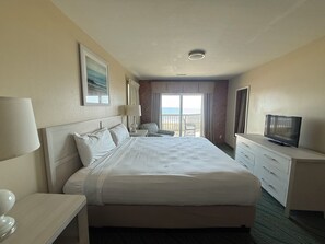 Quarto de hotel com uma cama grande, televisão, escrivaninha com abajur, cadeira e vista para o oceano pelas portas da varanda.