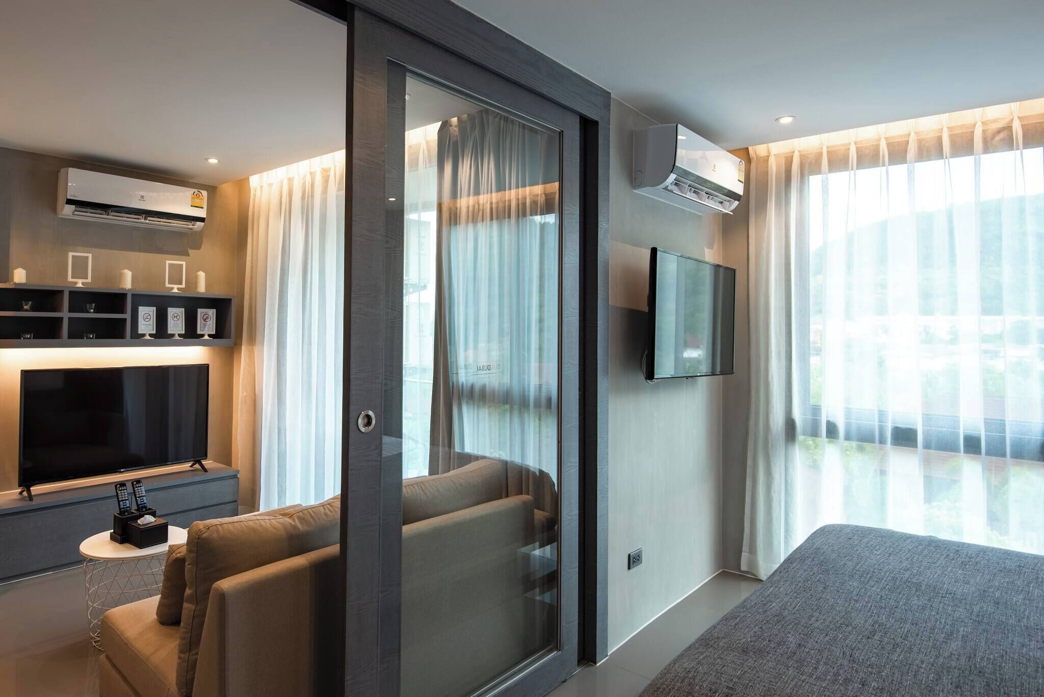 A modern hotel room with a sliding glass door, a flat-screen TV, a sofa, a bedside table with a remote control, and an air conditioning unit.