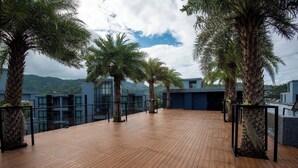 A wooden deck with palm trees, a glass railing, and a view of buildings and mountains. - Modern 1-bedroom Suite With Pool Access in Kamala, Phuket (Kamala)