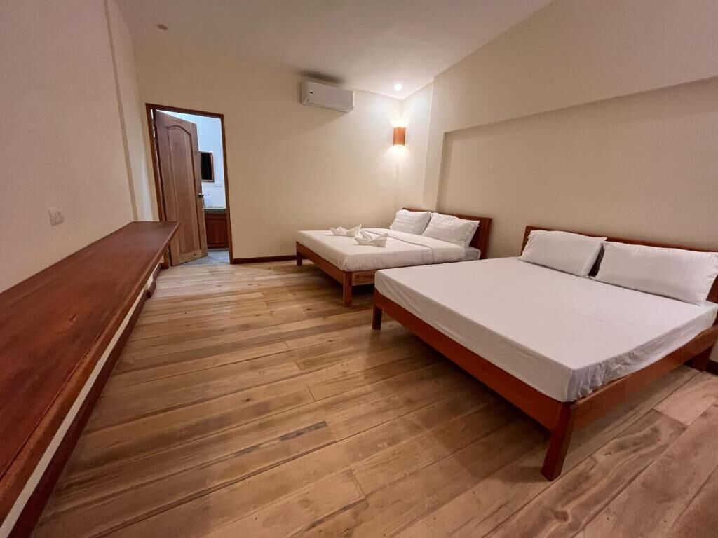 Una habitación con dos camas, suelo de madera, un banco y una lámpara fijada a la pared.