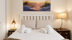 A neatly made bed with white linens, a painting of a sunset over a beach, a bedside lamp, and a tray with a book and a wooden object. - Private Retreat Near Interlochen Arts| Green Lake (Interlochen)