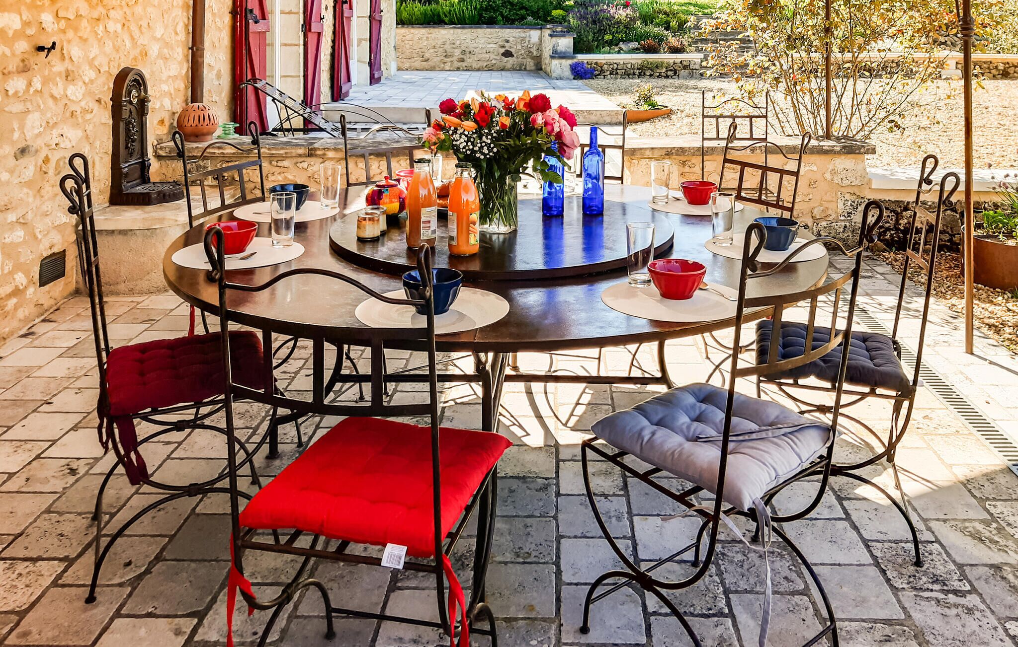 A round outdoor dining table with chairs, a vase of flowers, and various tableware.