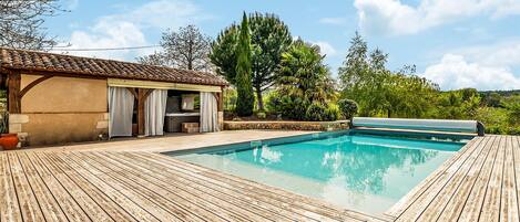 Una terraza de madera rodea una piscina rectangular con un pérgola y zona de estar al aire libre.