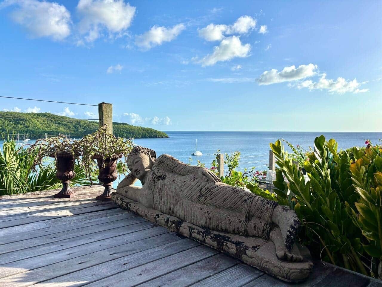 A wooden deck with a stone sculpture, potted plants, and a view of the sea and a distant island.