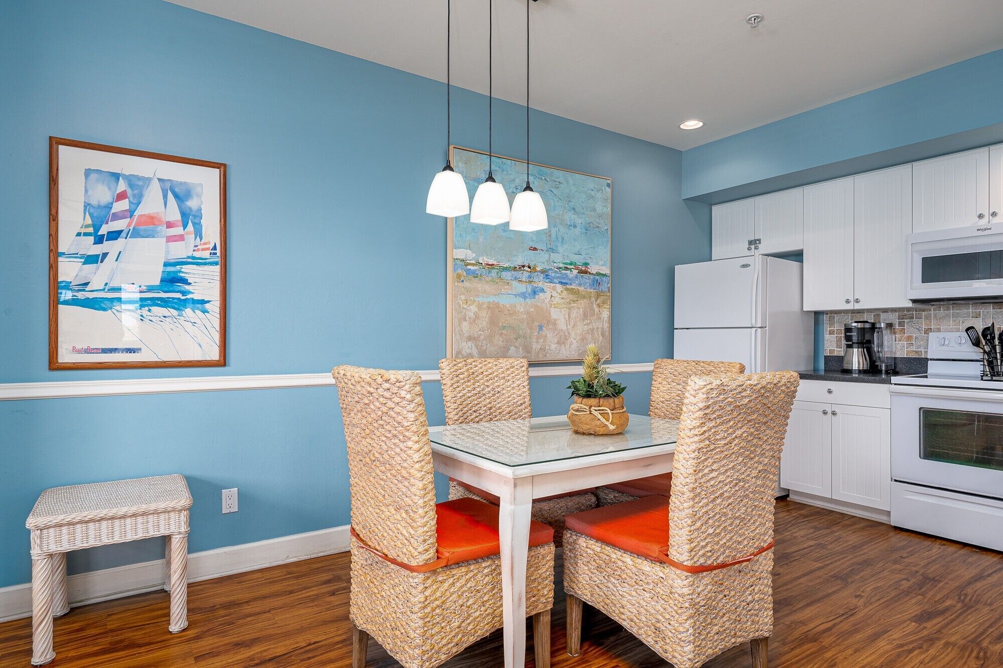 A dining area with wicker chairs and a glass table, a kitchen with white cabinets and appliances, and two paintings on the walls.