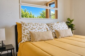 A bed with a yellow bedspread and floral pillows, a bedside table with a lamp, and a window with curtains. - 3 Bedroom | King Bed, 2 Queens | 5 Min to Kings Theatre, Near Trains and Cafes (Brookyn)