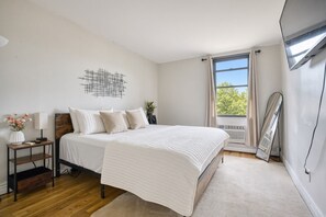 Un dormitorio con cama, mesitas de noche, una obra de arte en la pared, ventana con cortinas y un equipo de aire acondicionado.