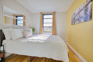 A bedroom with a bed, a mirror, a window with curtains, a plant, and a painting on the wall.