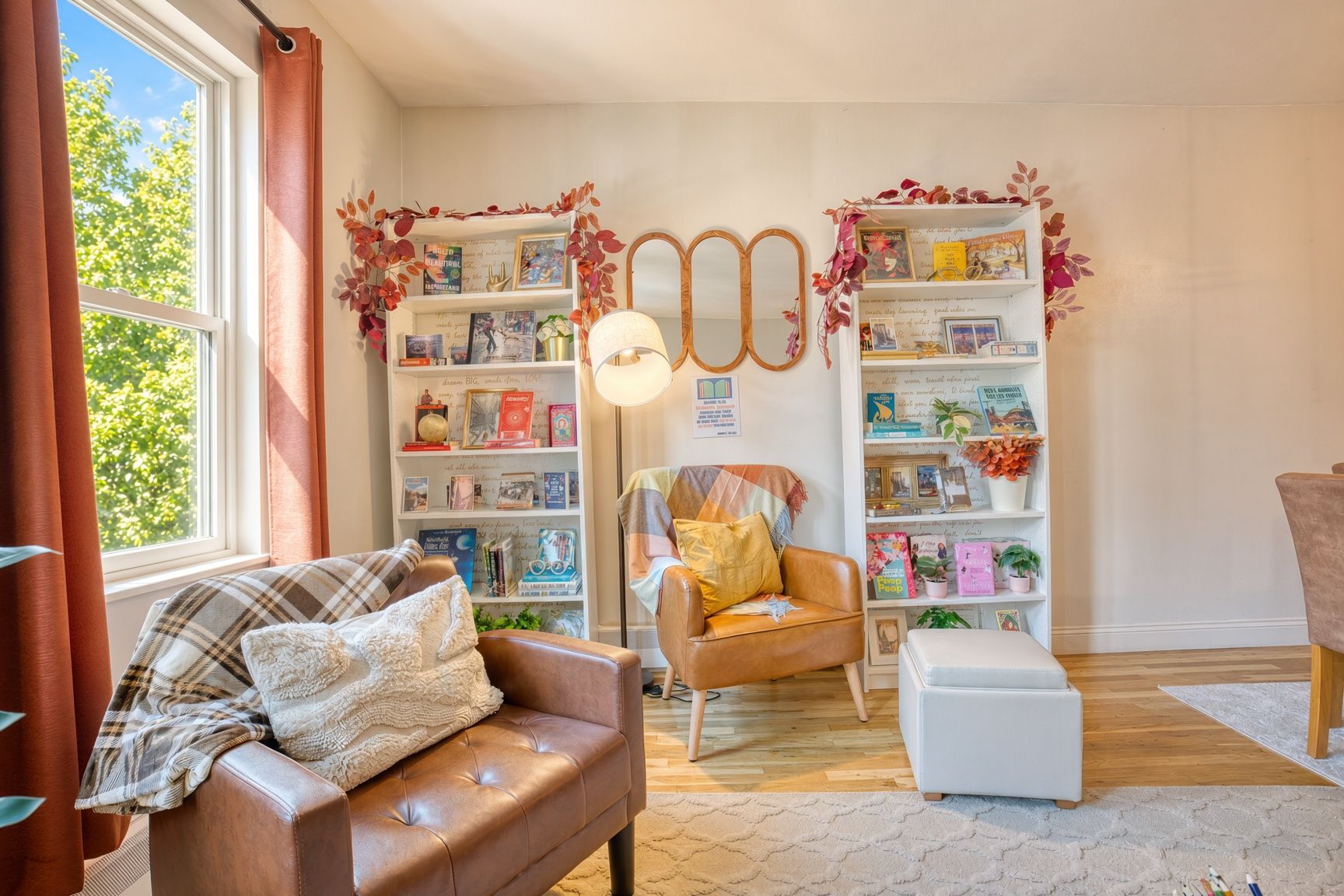 Una sala de estar acogedora con un sofá de cuero marrón, un reposapiés blanco y un sillón con un cojín amarillo. Hay dos estanterías blancas llenas de libros y objetos decorativos. Un espejo grande cuelga en la pared, y se ve una lámpara de pie.