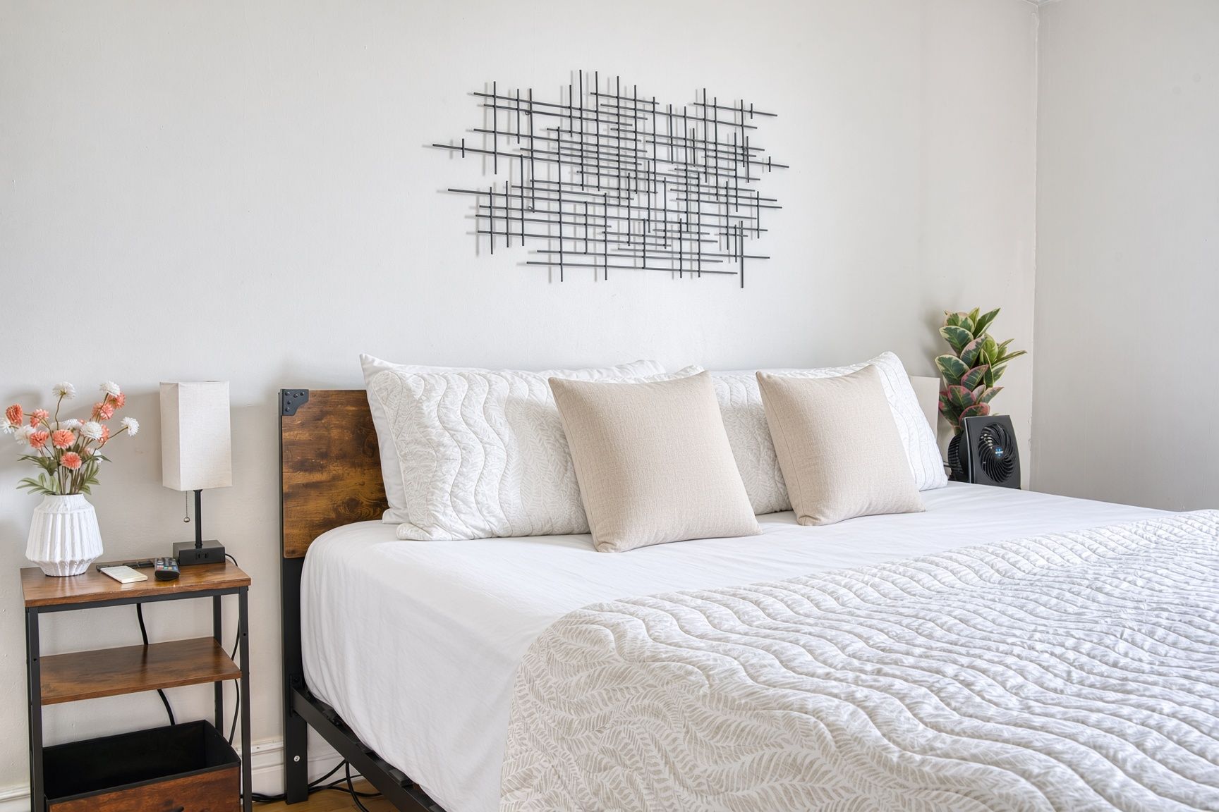 Un lit bien fait, recouvert d’une couette blanche, une table de chevet avec une lampe, une petite plante et une œuvre d’art accrochée au mur.