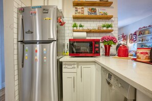 Una cocina con un refrigerador de acero inoxidable, un microondas rojo y un lavavajillas. Hay repisas de madera sobre la encimera con libros de cocina y un jarrón con flores.