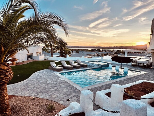 Zona de piscina con sillones reclinables, una palmera y vistas al atardecer.