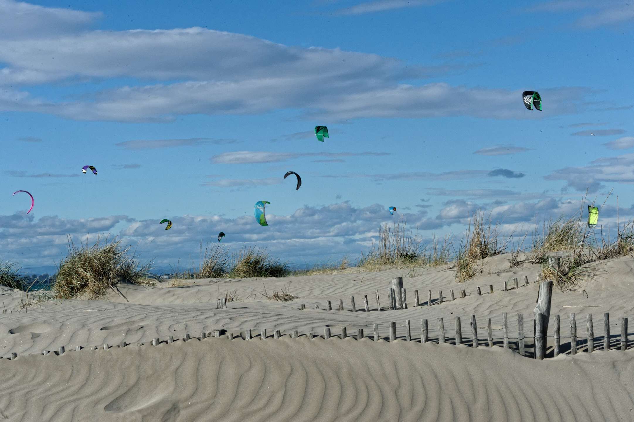 Una playa arenosa con una valla de madera y varios cometas volando en el cielo.