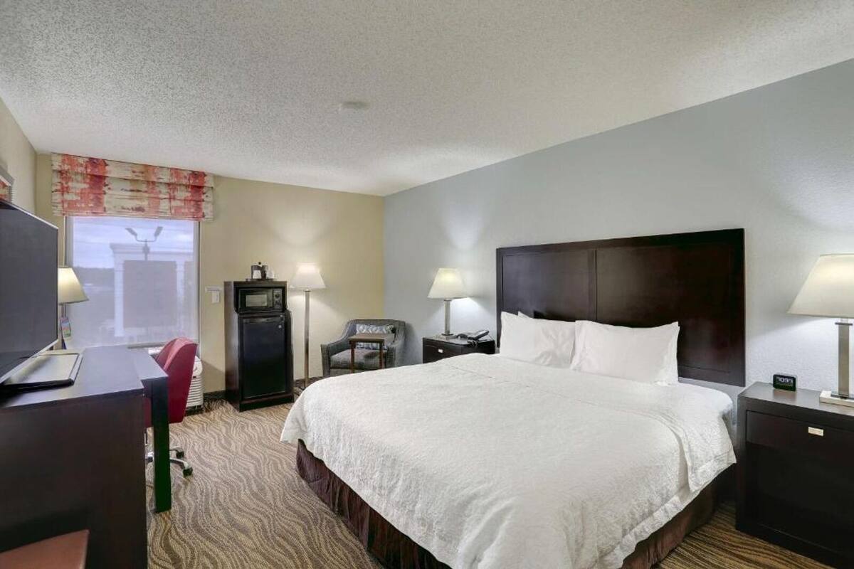 A hotel room with a large bed, a desk with a computer, a TV, a chair, a lamp, and a clock.