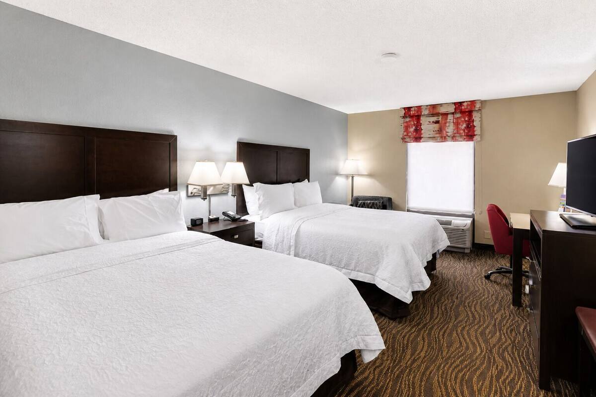 A hotel room with two beds, a desk, a chair, a TV, and a window with a red curtain.