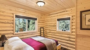 Une chambre à coucher aménagée dans un chalet en rondins, avec un lit, deux fenêtres, un luminaire au plafond et un tableau encadré accroché au mur.