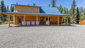 Une cabane en rondins dotée d’un porche couvert, d’une allée en gravier et bordée par une forêt.
