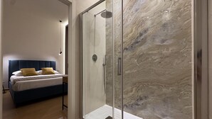 Quarto moderno com cabine de chuveiro de vidro, cama com cabeceira azul e piso de madeira.
