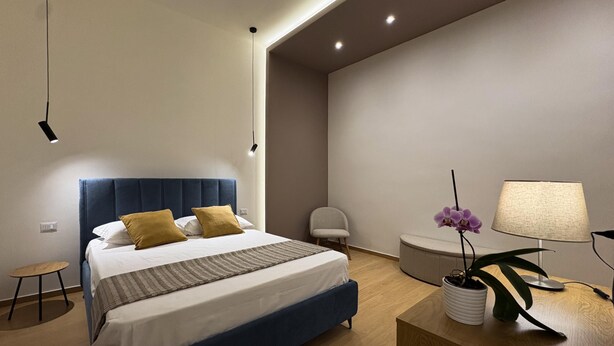 Quarto moderno com cama, mesa de cabeceira, luminária, cadeira e uma pequena mesa com uma planta.