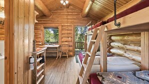 A log cabin interior with a bunk bed, a dining area with a table and chairs, a window with a view of greenery, and a ceiling fan.