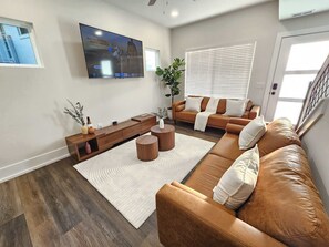 Ein Wohnzimmer mit braunem Ledersofa, einem Holztisch, einem an der Wand montierten Fernseher und einem Fenster mit Jalousien.