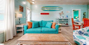 A room with a teal sofa, a wooden coffee table, a surfboard, and a beach-themed chair. - SP3 Shared Pool, Pet Friendly, Parking for 3 (Port Aransas)