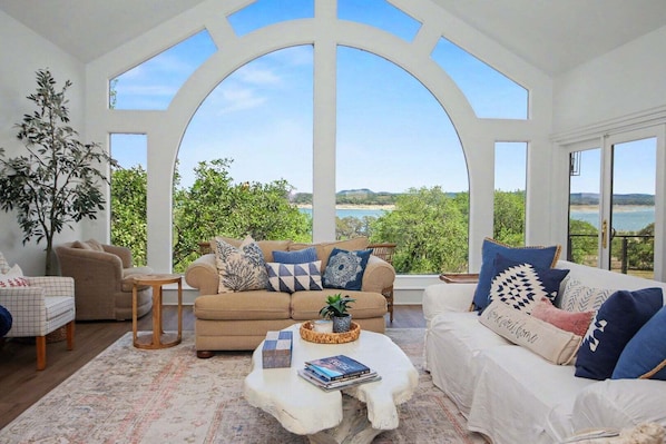 A spacious living room with a large arched window, a beige sofa, a white sofa, and a coffee table with books and a plant. - Meet Me In Paradise: CL Waterfront + Outdoor Fireplace + Pool Table + Ping Pong + 2 Living Areas (Canyon Lake)