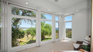 A spacious room with large windows, a wicker chair, and a view of a garden and water. - Meet Me In Paradise: CL Waterfront + Outdoor Fireplace + Pool Table + Ping Pong + 2 Living Areas (Canyon Lake)