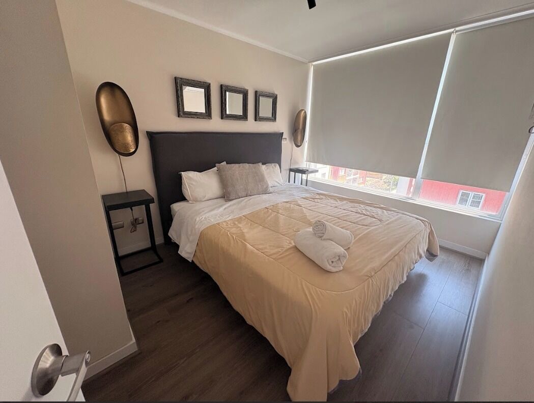 A bedroom with a bed, bedside tables, a window with blinds, and a view of buildings outside.