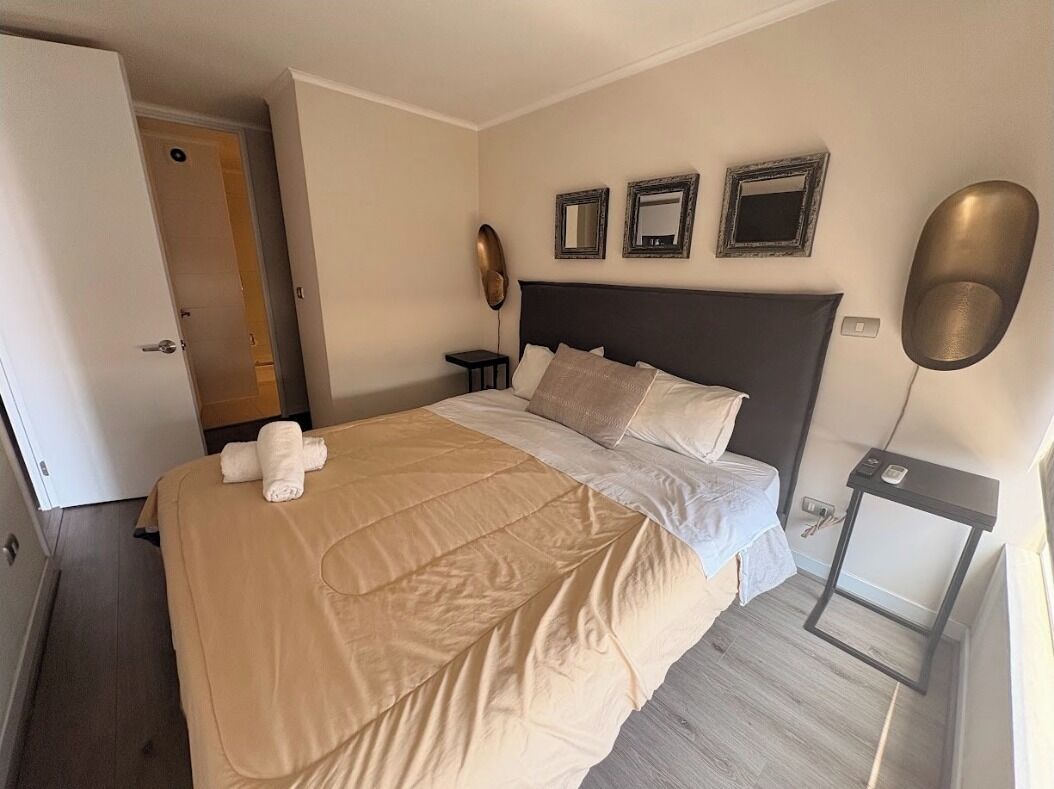 A neatly made bed with a beige blanket, a bedside table with a lamp, and three framed pictures on the wall.