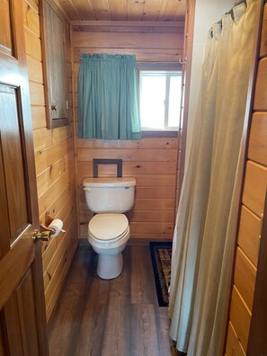 A small, wooden cabin bathroom with a toilet, a window with a curtain, and a door. - Amazing 4-bedroom cabin in Brownville perfect for your getaway (Brownville)