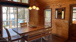 A wooden cabin interior with a dining table, chairs, a chandelier, and large windows overlooking a forested area. - Amazing 4-bedroom cabin in Brownville perfect for your getaway (Brownville)