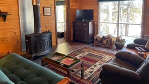 A cozy cabin interior with a wood-burning stove, a television, and a large window offering a view of trees. - Amazing 4-bedroom cabin in Brownville perfect for your getaway (Brownville)