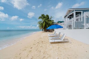 A beach with white lounge chairs, a blue umbrella, and a white building with a balcony. - Beachfront Villa with Plunge Pool - Sea Breeze Beach Villa (Fitts Village)