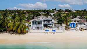 Ein Strandgrundstück mit einem zweigeschossigen Haus, einem Balkon sowie einem Strandbereich mit Liegestühlen und Sonnenschirmen.