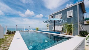 Ein Swimmingpool mit einem blau-weiß gestreiften Sonnenschirm, einer weißen Liegefläche und einem Haus mit weißen Fensterläden.