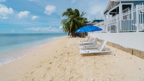 A beach with white lounge chairs, a blue umbrella, and a building with a white fence. - Beachfront Villa with Plunge Pool - Sea Breeze House (4 bed) (Fitts Village)