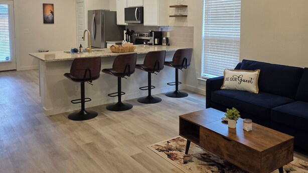 A modern kitchen with a central island, bar stools, a dark sofa, and a ceiling fan. - New 3-bedroom, 2 bath, 2 car garage, home in exceptional south Lubbock location. (Lubbock)
