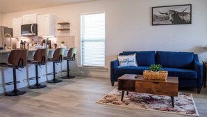 A modern living room with a blue sofa, a coffee table, and a kitchen area with white cabinets and a bar stool setup. - New 3-bedroom, 2 bath, 2 car garage, home in exceptional south Lubbock location. (Lubbock)
